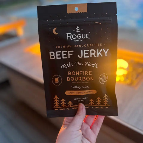 Holding a bag of Rogue Bonfire Bourbon Beef Jerky with a fire in the background.