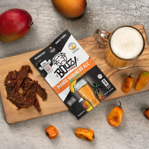 A bag of Boozy Mango Habanero Ale jerky on a cutting board next to a mug of craft beer, with beef jerky and habanero peppers surrounding it.
