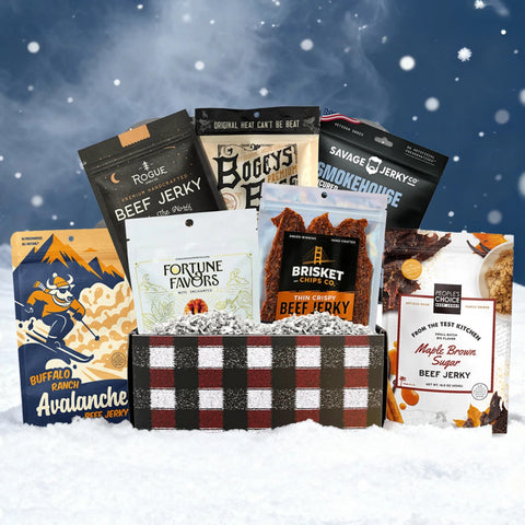 JerkyGent Holiday Edition plaid gift box displayed in the snow, filled with assorted small-batch beef jerky bags and Fortune Favors pecans against a snowy winter background.