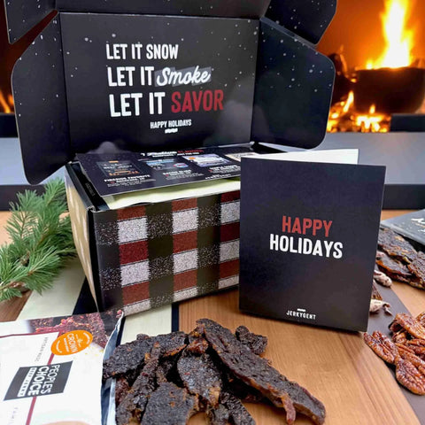 Open JerkyGent Holiday Gift Box with plaid design, showing festive messaging “Let it Snow, Let it Smoke, Let it Savor” inside the lid. A black “Happy Holidays” greeting card sits in front alongside jerky pieces on a wooden table.