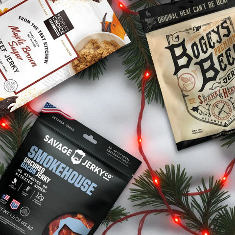 Assorted jerky bags from Savage Jerky, People’s Choice, and Bogey’s Beef on snowy background with pine and holiday lights.