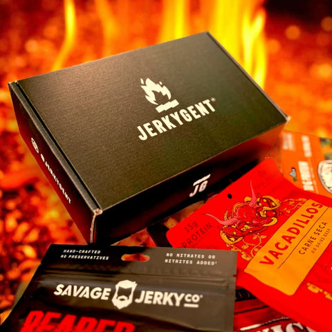 The JerkyGent Inferno Box surrounded by assorted spicy craft jerky packs, set against a backdrop of fire.