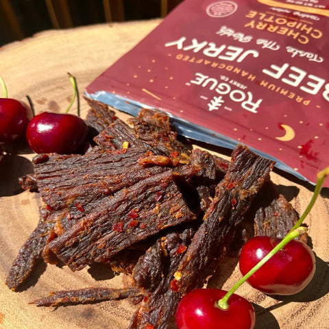 Cherry beef jerky and fresh cherries on a wooden board with Rogue Jerky packaging.
