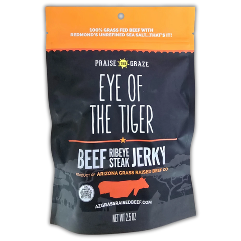Front of a “Praise the Graze – Eye of the Tiger” Beef Ribeye Steak Jerky bag. The design features bold white and orange text on a black matte background. The top reads “100% Grass Fed Beef with Redmond’s Unrefined Sea Salt…That’s It!” Below, the name “Eye of the Tiger” is prominently displayed with “Beef Ribeye Steak Jerky” underneath. It is labeled as a product of Arizona Grass Raised Beef Co., with a red cow silhouette and mountain range graphics at the bottom. The net weight is 2.5 oz.