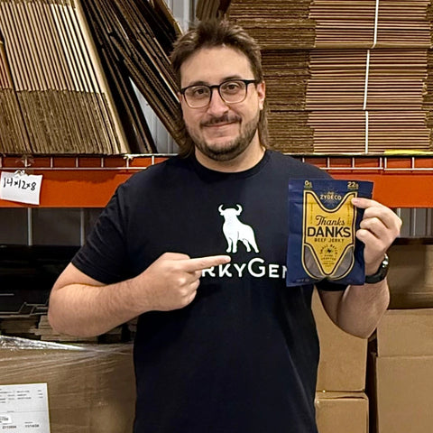 Jeff from JerkyGent pointing at a pack of Thanks Danks Zydeco Beef Jerky, standing in front of orange warehouse shelves.