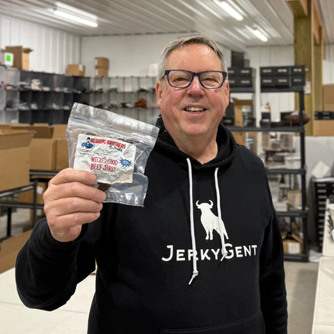 John holding Herring Brothers’ Wicked Good Beef Jerky and smiling in the JerkyGent packing area, wearing a branded hoodie.