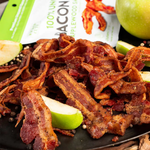 Close-up of Wicked Cutz Applewood Smoke Bacon Jerky with green apple slices and whole peppercorns, showcasing crispy uncured bacon pieces in front of product packaging.
