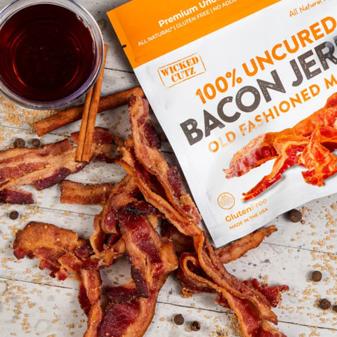 Wicked Cutz Old Fashioned Maple Bacon Jerky on a rustic surface with brown sugar, cinnamon sticks, and a glass of maple syrup, with the product bag partially visible.