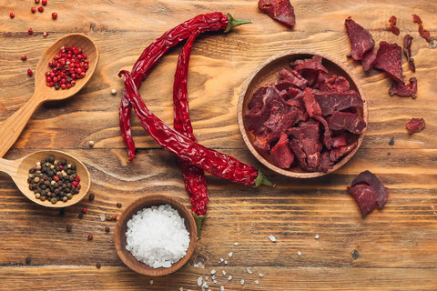 A top-view of a tabletop with a bowl of spicy beef jerky, red Chile peppers, salt, and other spices.