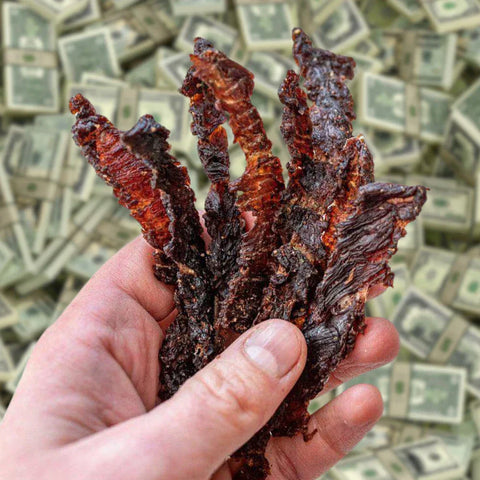 A handful of delicious beef jerky being held over a large pile of US currency.