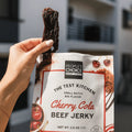 Hand holding a piece of cherry cola beef jerky above its packaging.