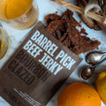Double Neat Bourbon Glazed beef jerky next to a glass of whiskey and oranges.