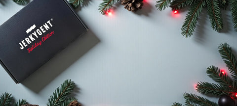 JerkyGent Holiday Gift Box on a snowy backdrop with red christmas lights and fir branches.