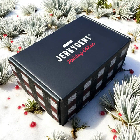 Black JerkyGent Holiday Edition gift box with plaid design, displayed on snow with frosted pine and red berries.