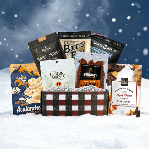 JerkyGent Holiday Edition plaid gift box displayed in the snow, filled with assorted small-batch beef jerky bags and Fortune Favors pecans against a snowy winter background.