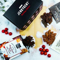 JerkyGent Holiday Edition gift box surrounded by Maple Brown Sugar jerky, Rogue Jerky, Fortune Favors candied pecans, and festive red ornaments on a snowy wooden surface.