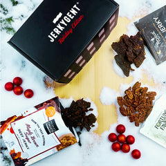 JerkyGent Holiday Edition gift box surrounded by Maple Brown Sugar jerky, Rogue Jerky, Fortune Favors candied pecans, and festive red ornaments on a snowy wooden surface.