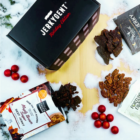 JerkyGent Holiday Edition gift box surrounded by Maple Brown Sugar jerky, Rogue Jerky, Fortune Favors candied pecans, and festive red ornaments on a snowy wooden surface.
