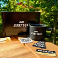 JerkyGent black mug, branded gift box, and sticker set displayed outdoors on a wooden surface in the sunlight.