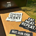 Close-up of JerkyGent stickers on a wooden table, featuring “Powered by Jerky,” “Eat. Meat. Repeat.” and “Best. Club. Ever.” designs.