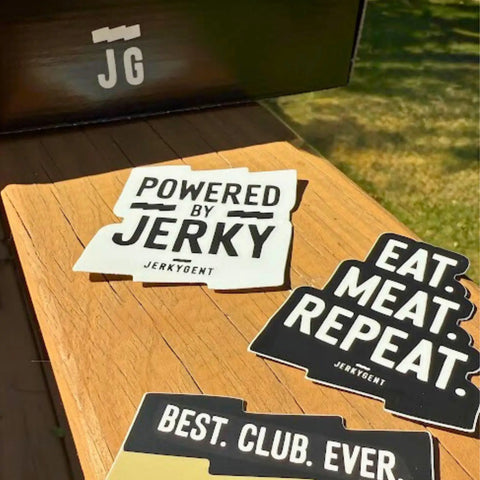 Close-up of JerkyGent stickers on a wooden table, featuring “Powered by Jerky,” “Eat. Meat. Repeat.” and “Best. Club. Ever.” designs.