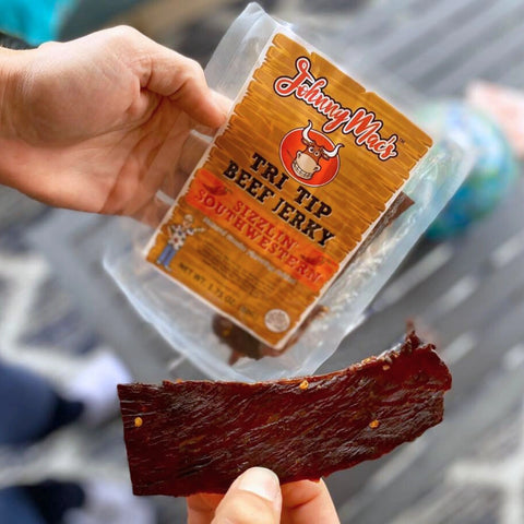 A close-up photo of someone holding the packaging and a piece of Johnny Mac's Sizzlin' Southwestern Tri-Tip beef jerky 