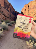 A hand holds a bag of Mojave Jerky Co. Beef Jerky in front of a desert canyon landscape with red rock cliffs and a dirt path.