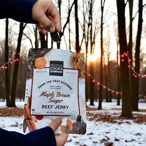 Hand holding a bag of People’s Choice Maple Brown Sugar beef jerky in a snowy forest at sunset with red holiday string lights.