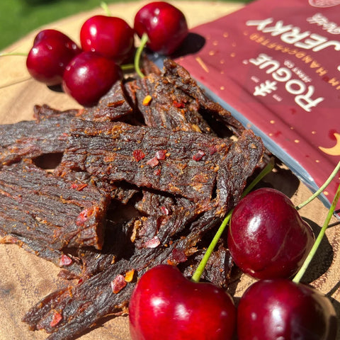 A close-up photo of Rogue Jerky Co. Cherry Chipotle beef jerky along with several Traverse City, MI cherries