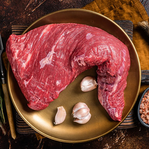 A large raw tri-tip steak sitting in a plate with some garlic cloves