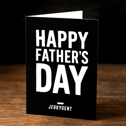 A black Father's Day greeting card with bold white text that reads "HAPPY FATHER'S DAY" on the front. The JerkyGent logo appears at the bottom. The card is standing upright on a wooden surface against a dark background.