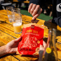 A person shares a piece of People's Choice Carne Seca Chipotle & Roasted Garlic Beef Jerky at an outdoor gathering. The bold red bag sits on a rustic wooden table with glasses of beer, showcasing this jerky as the perfect snack for social occasions.