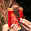 A close-up of a hand holding a piece of People's Choice Carne Seca Chipotle & Roasted Garlic Beef Jerky, highlighting its textured, handcrafted appearance. The red bag is blurred in the background, focusing on the jerky's quality and rich seasoning.