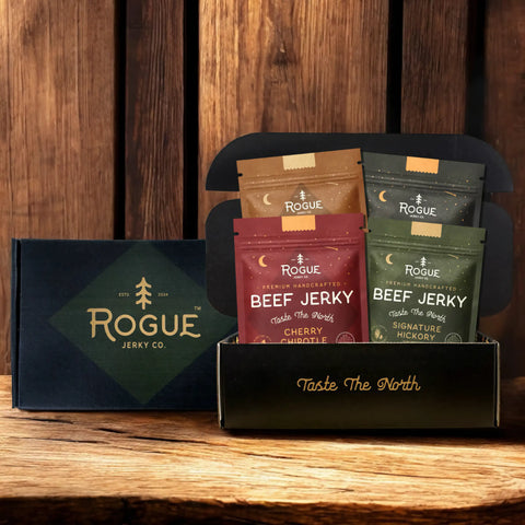 Rogue Jerky Co. beef jerky gift box featuring four premium handcrafted flavors—Cherry Chipotle, Maple & Brown Sugar, Signature Hickory, and Bonfire Bourbon—packaged in a rustic gift box with 'Taste The North' branding against a wood backdrop.